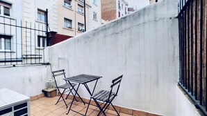 Une petite terrasse extérieure avec une table en métal et deux chaises, un mur blanc et une clôture en métal.