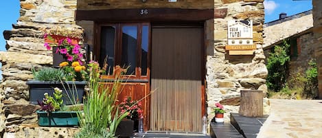 A rustic stone house with a wooden balcony, a wooden door, and potted plants at the entrance.