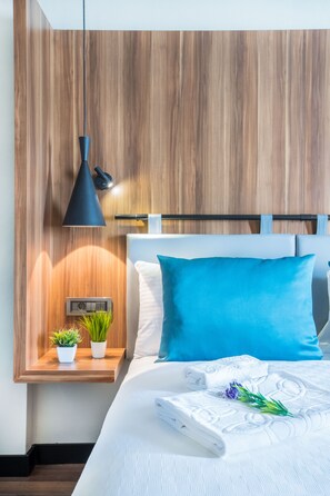 A neatly made bed with white bedding and a blue pillow, a bedside table with a small plant, a wall-mounted light fixture, and a wooden panel wall. - Signature Hotels & Spa (Ürgüp)