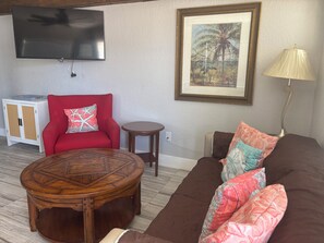 A living room with a red armchair, a brown sofa, a wooden coffee table, a small round table, a lamp, a television, and a framed picture on the wall.