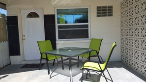 Een terras met een glazen tafel en vier stoelen, een witte deur en een raam met jaloezieën.