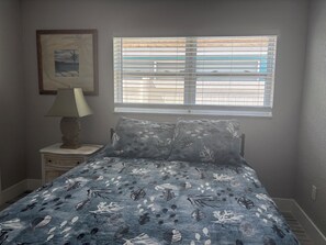 A bedroom with a bed, a nightstand, a lamp, a window with blinds, and a framed picture on the wall.