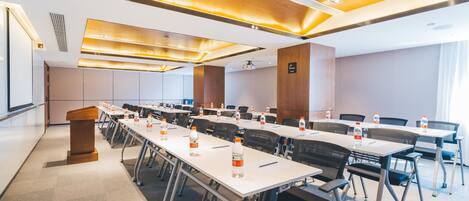 A conference room with multiple tables and chairs, equipped with water bottles, a podium, and a projector screen.
