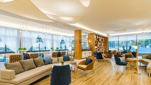 Un hall d’hôtel moderne, doté d’un aménagement spacieux, de grandes fenêtres et de divers types de sièges, notamment des canapés et des fauteuils.