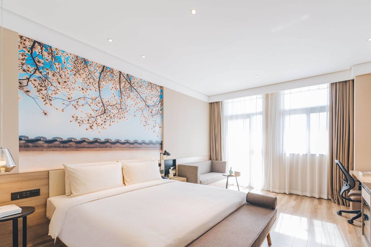 A modern hotel room with a large bed, a desk, a chair, and a large window with curtains.