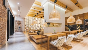 A modern interior with a long hallway leading to a seating area with tables and chairs, wooden beams on the ceiling, and a brick wall on one side.