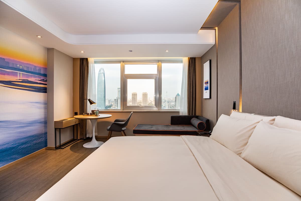 A modern hotel room with a large bed, a desk with a chair, a small table, and a view of the cityscape through the window.