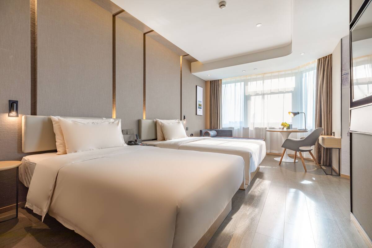 A hotel room with two beds, a desk, a chair, and a large window with curtains.