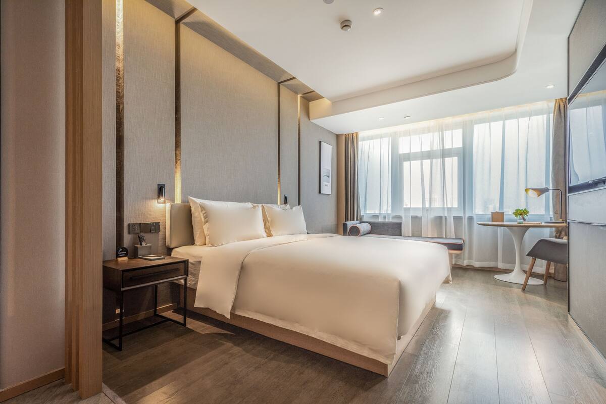 A modern hotel room with a large bed, bedside tables, a desk, and a view of the city through large windows.