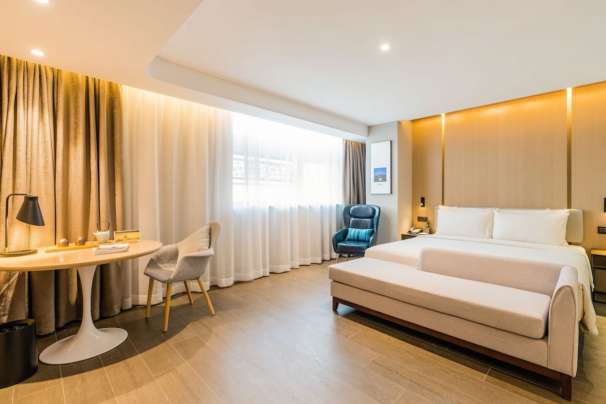 Quarto de hotel com cama, escrivaninha com luminária, cadeira e uma janela grande com cortinas.
