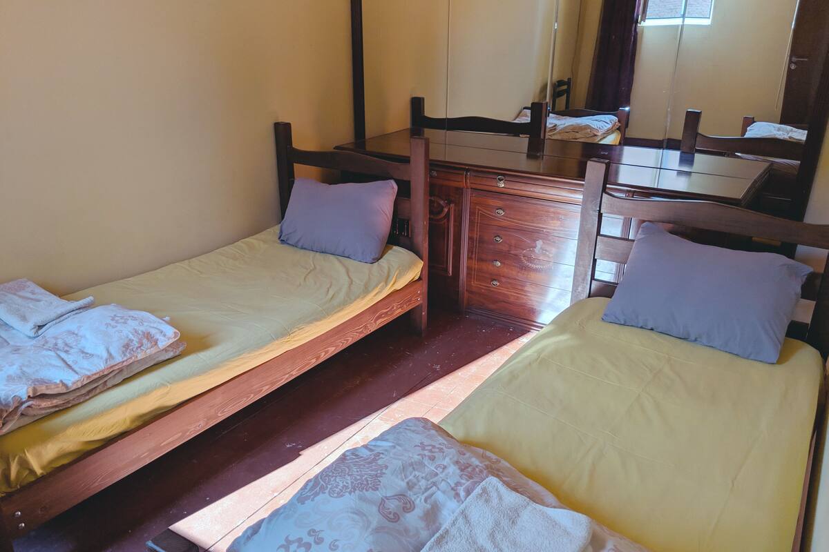 A room with two wooden beds, each with a pillow and a blanket. There is a wooden dresser with drawers and a mirror above it.