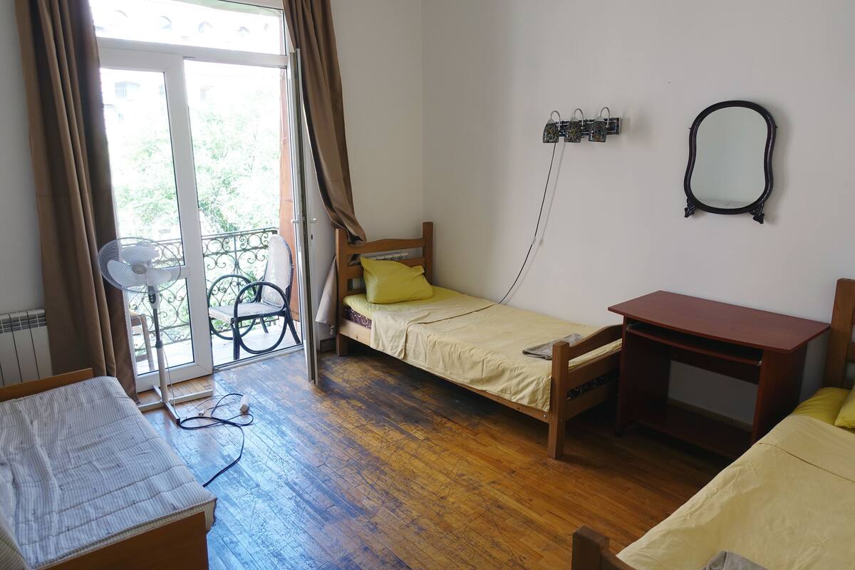 A small, single-room accommodation with a wooden floor, a bed with a yellow pillow, a wooden desk, a small table, and a wall-mounted mirror.