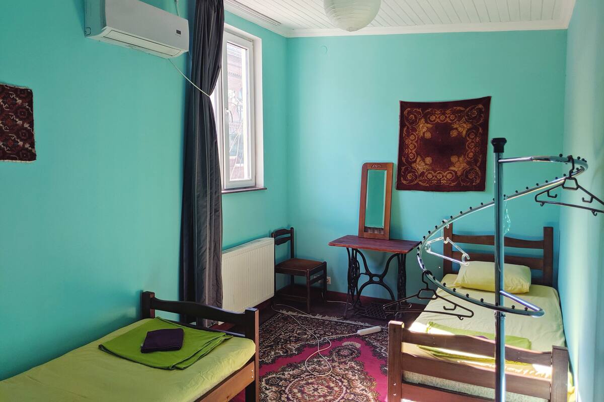 A room with teal walls, a bed with a green mattress, a wooden chair, a radiator, a mirror, a hanging light fixture, and a window with curtains.
