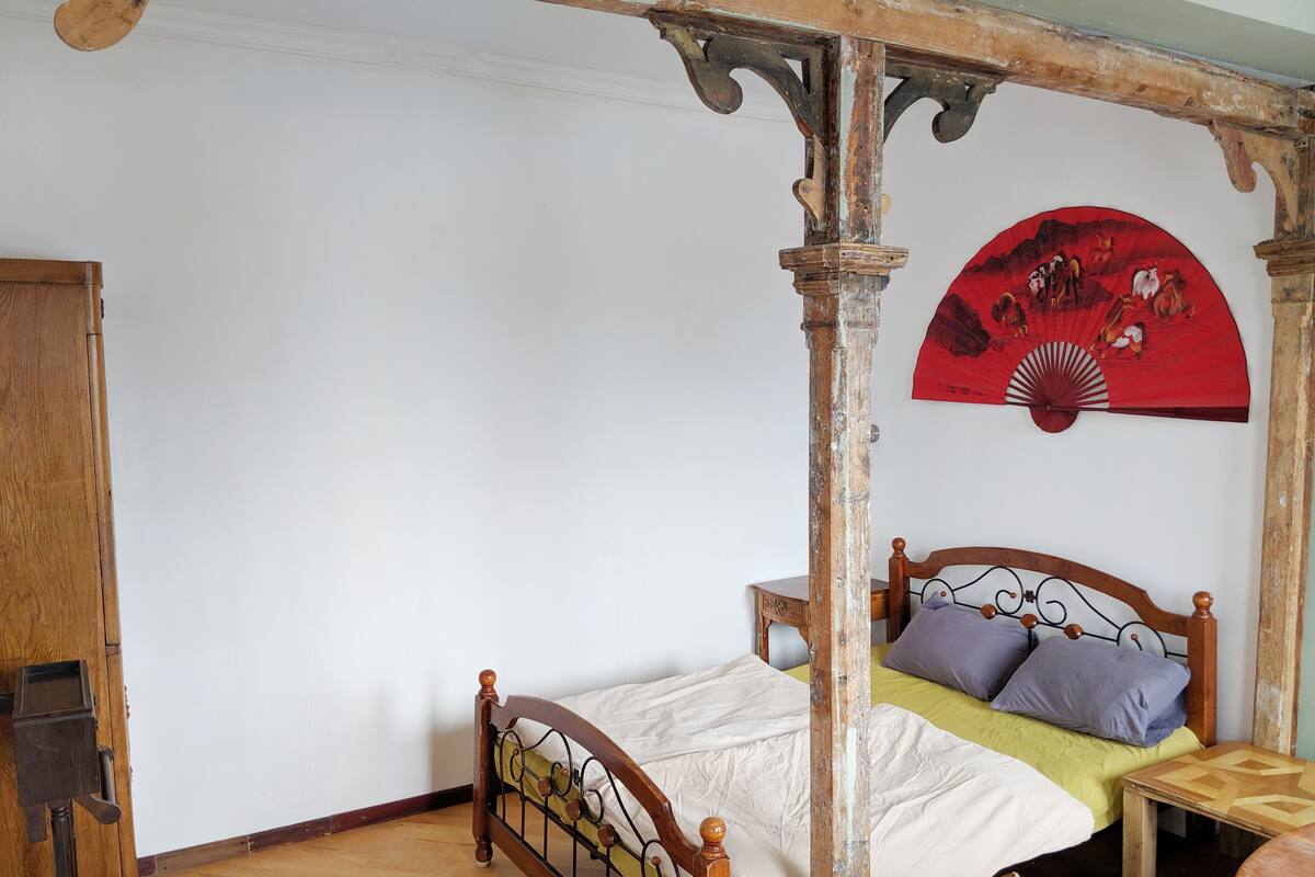 A four-poster bed with a wooden frame, a single bed with a yellow bedspread, a wooden nightstand, and a red decorative fan on the wall.