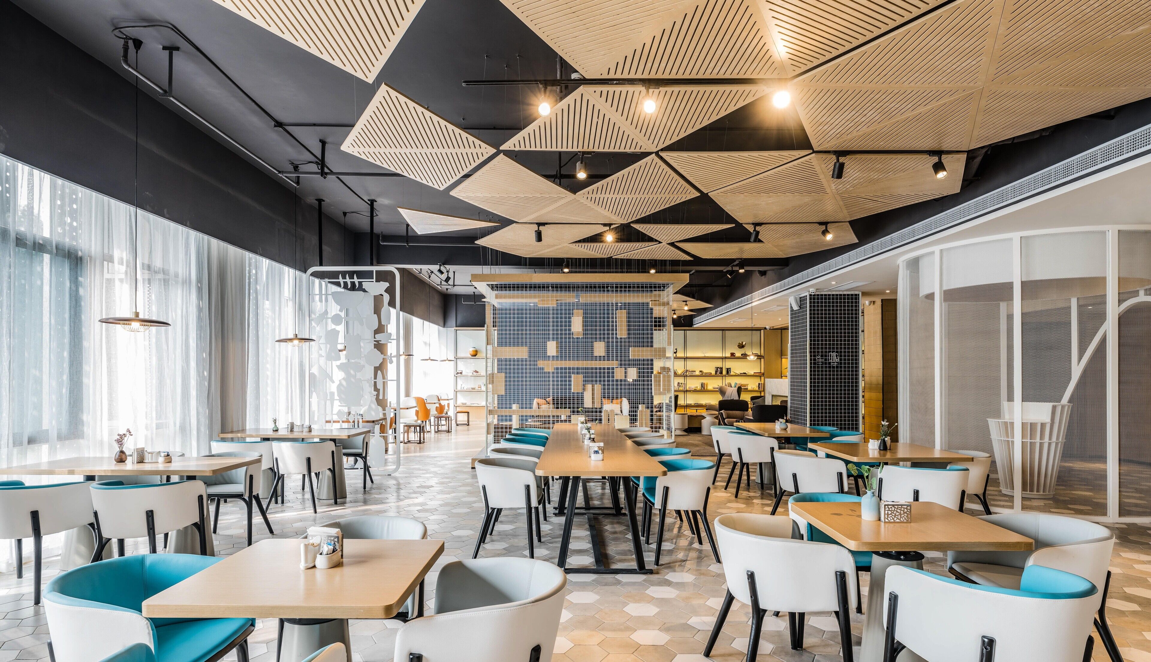 A modern restaurant with a long communal table, individual seating, and a patterned ceiling.