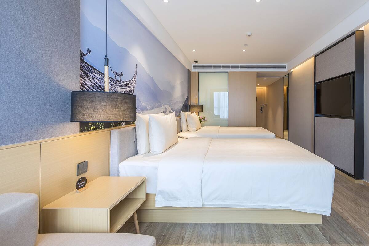 A hotel room with two beds, a desk, a TV, and a wall-mounted lamp.