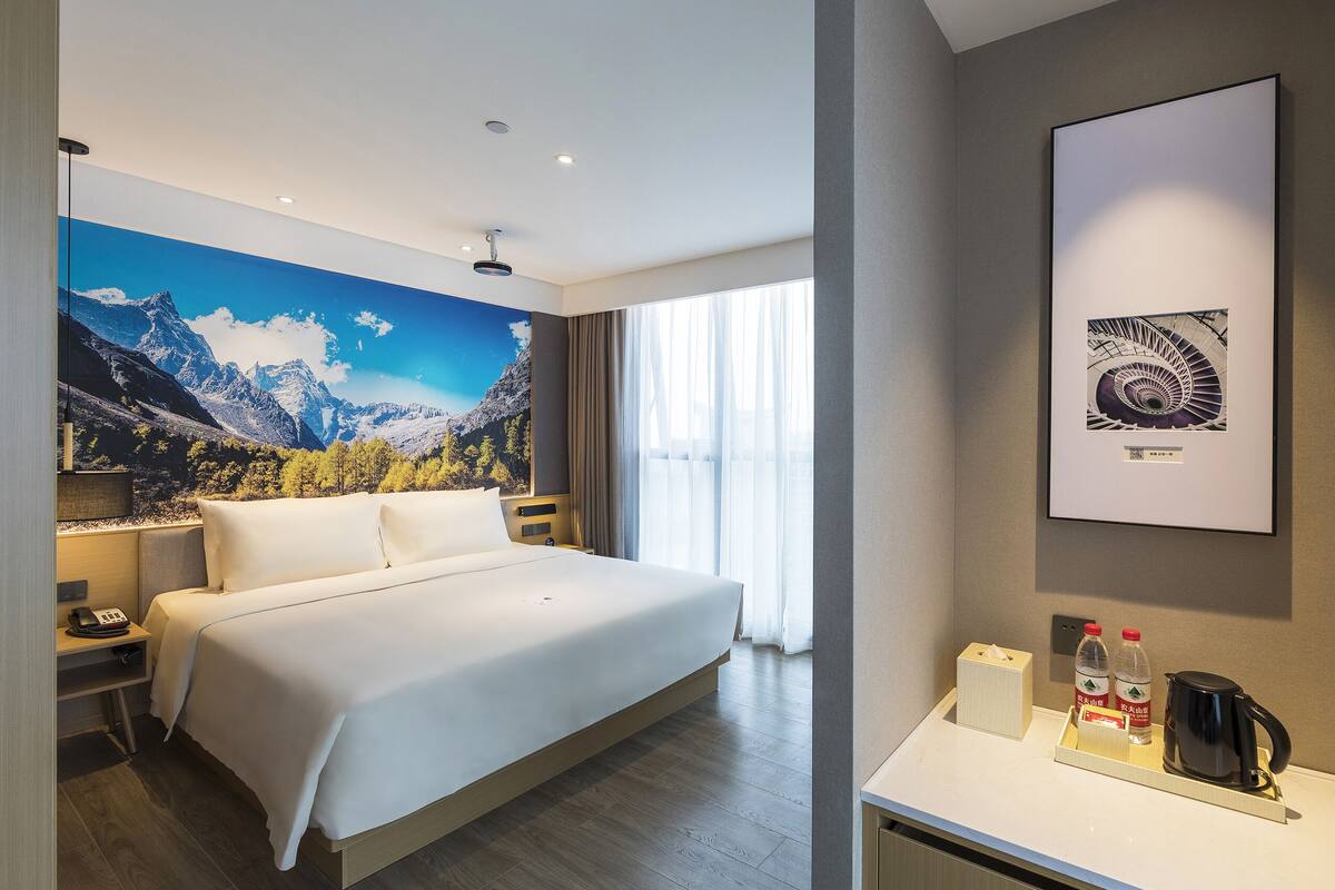 A hotel room with a bed, a desk with a coffee maker, a framed picture, and a window with curtains.