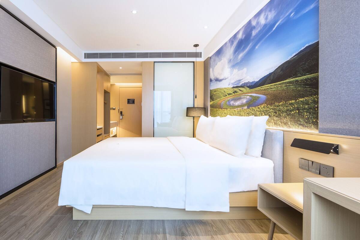 A hotel room with a large bed, a flat-screen TV, a painting of a landscape, and a desk with a lamp.
