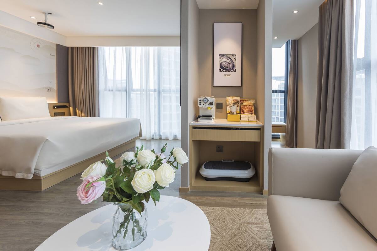 A hotel room with a bed, a desk with a sewing machine, a chair, a coffee table with a vase of flowers, and a large window with curtains.