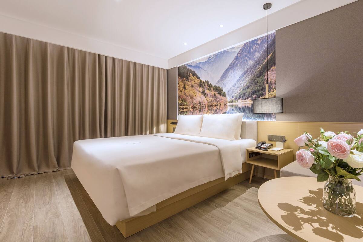 A hotel room with a large bed, a bedside table with a phone and a lamp, a small table with a vase of flowers, and a scenic mountain view on the wall.