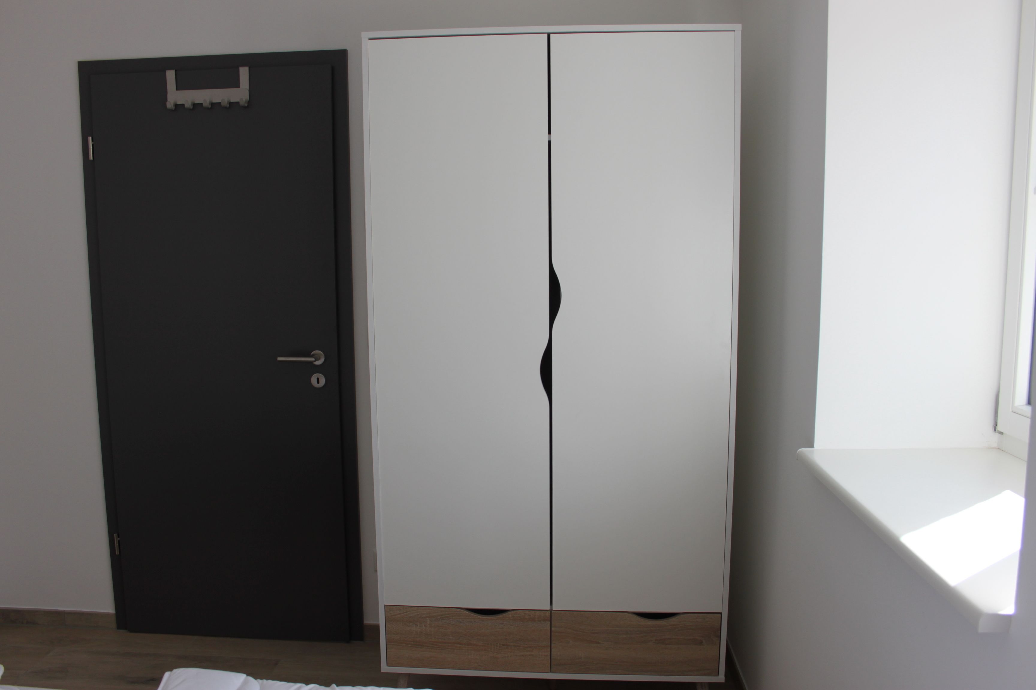 A room with a black door, a white wardrobe with a mirror, and a window.