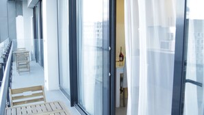 Ein Balkon mit Holztischen und -stühlen, große Fenster mit leichten Vorhängen und ein Blick in einen Raum, in dem sich eine Flasche auf einer Theke befindet.