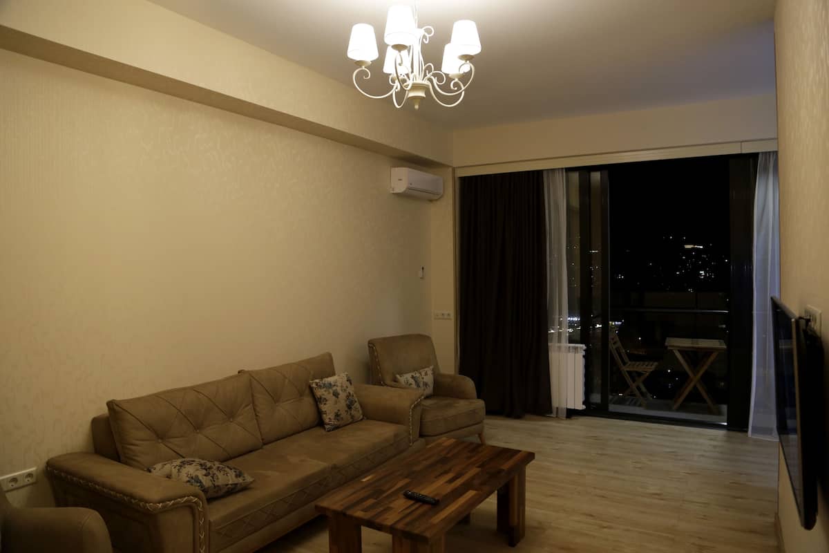 A living room with a sofa, a coffee table, and a chandelier. There is a window with curtains and a view of the city at night.
