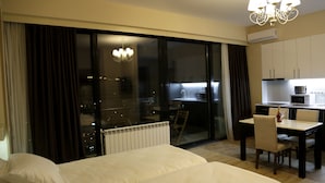 Une chambre d’hôtel avec un grand lit, une table à manger avec des chaises, un lustre, une vue à travers une porte vitrée et un coin cuisine.
