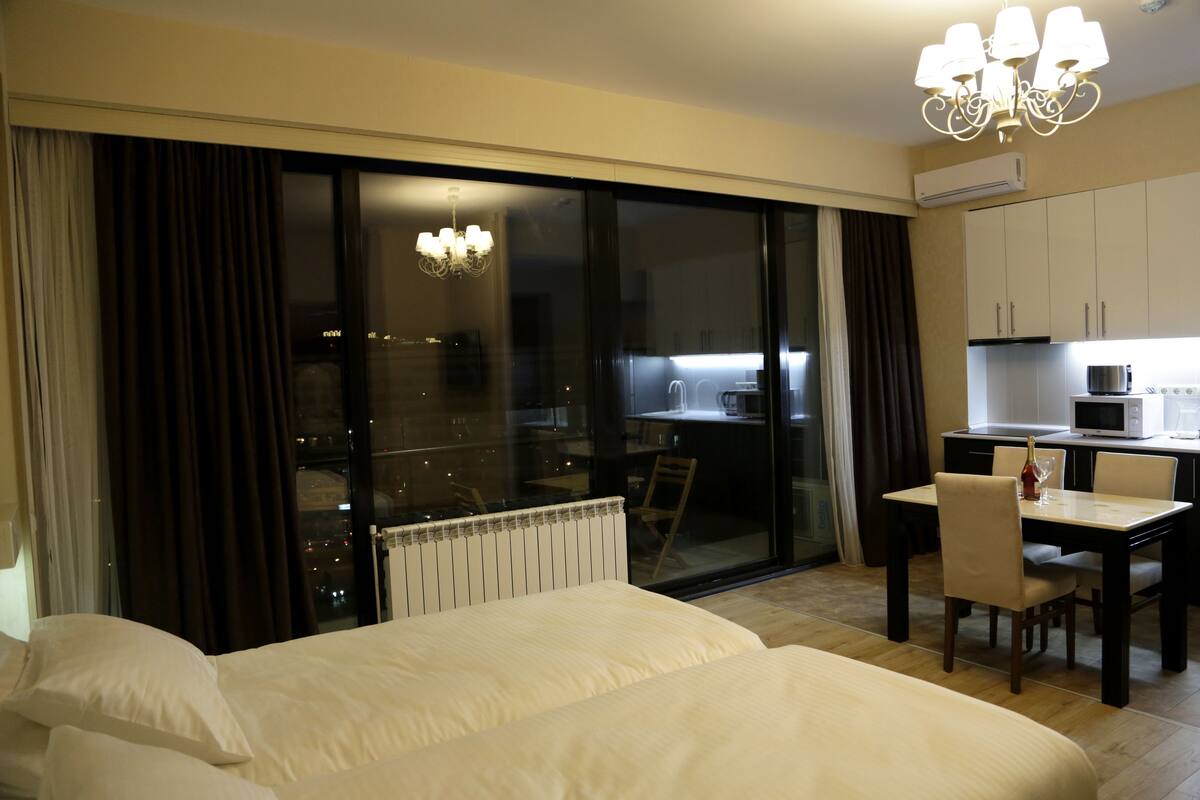 A hotel room with a large bed, a dining table with chairs, a chandelier, a view through a glass door, and a kitchenette area.