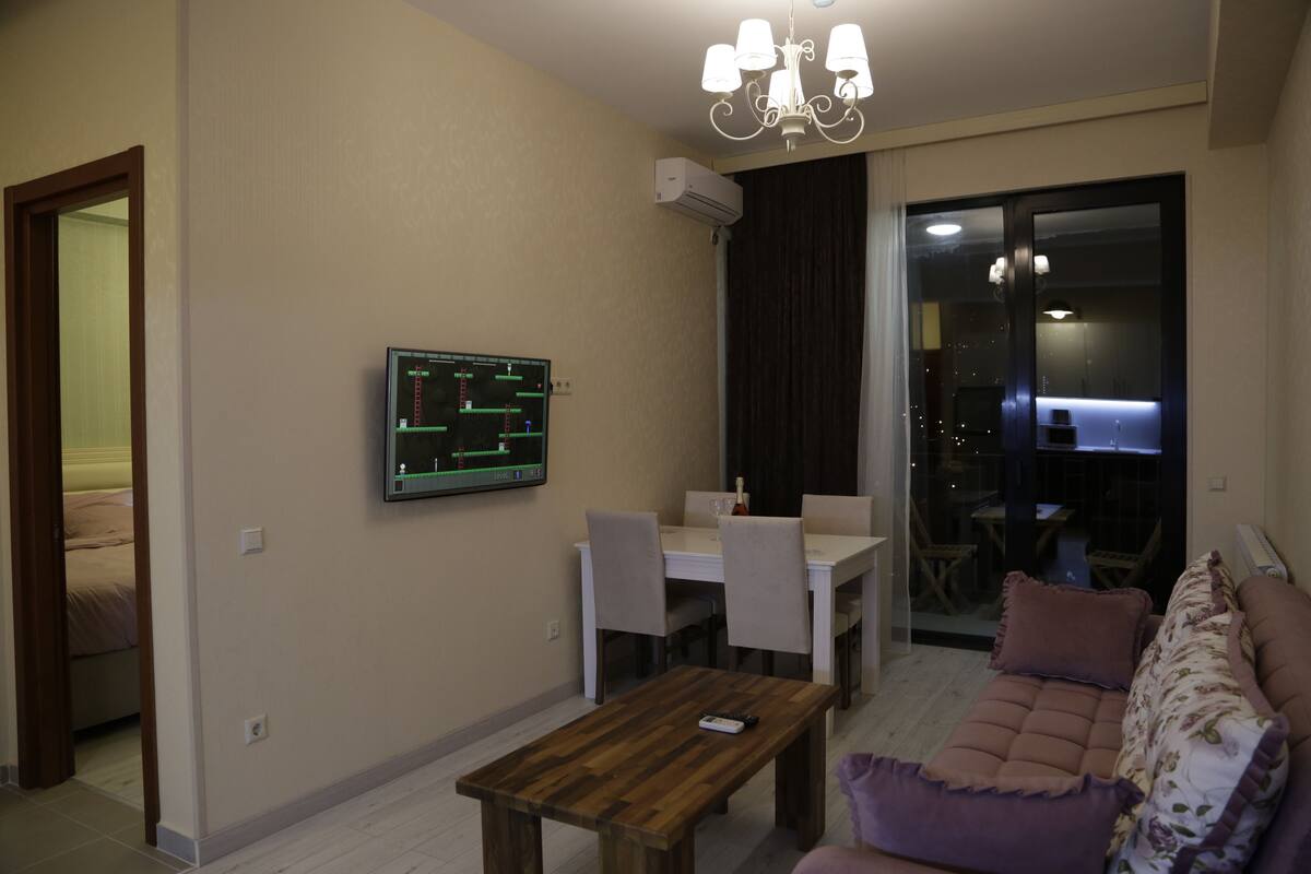 A living room with a wooden coffee table, a sofa, a dining table with chairs, and a TV mounted on the wall.