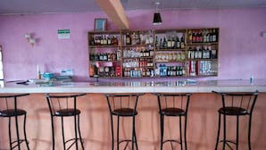 A bar with a long counter and four stools. Shelves behind the counter hold various bottles and glasses. - Gee -Thorn Hotel (Chaka)