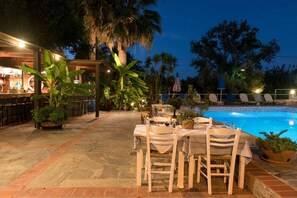 An outdoor pool area with tables and chairs, surrounded by tropical plants and a bar area.