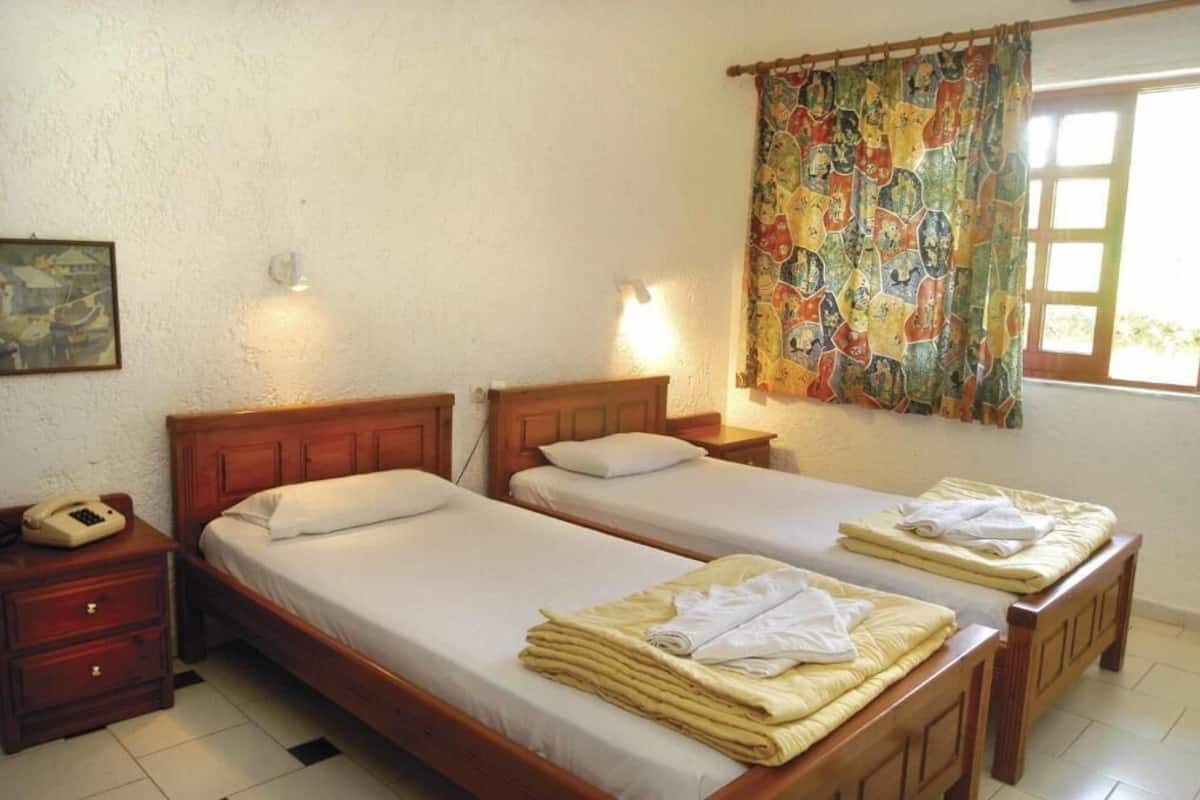 Um quarto com duas camas de madeira, uma mesa de cabeceira com telefone e uma tapeçaria colorida na janela.