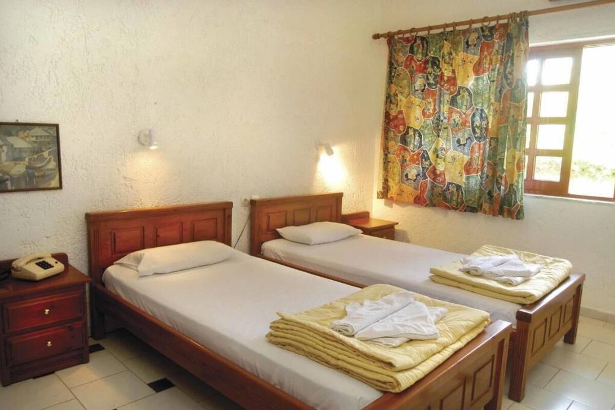 Um quarto com duas camas de madeira, uma mesa de cabeceira com telefone e uma tapeçaria colorida na janela.