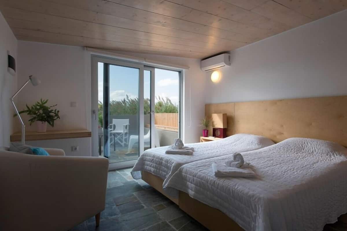 Quarto de hotel com duas camas, uma escrivaninha, uma cadeira, um vaso de planta e uma porta de vidro deslizante para uma área externa.