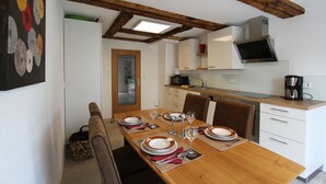 Un coin repas avec une table en bois dressée pour quatre personnes, une cuisine équipée d’appareils modernes et une grande fenêtre au-dessus de la table.