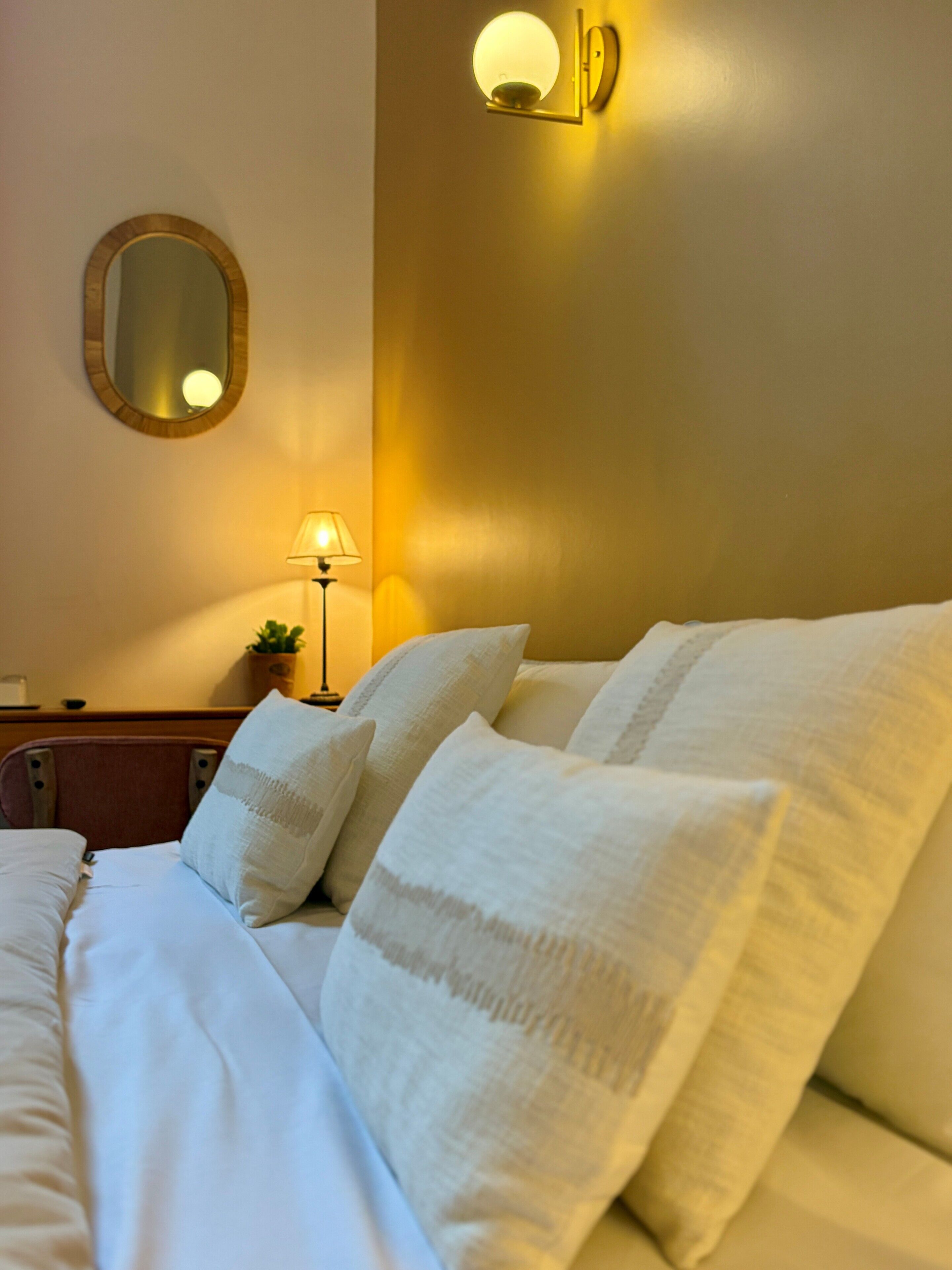A neatly made bed with white linens and pillows, a round mirror on the wall, and a wall-mounted light fixture.