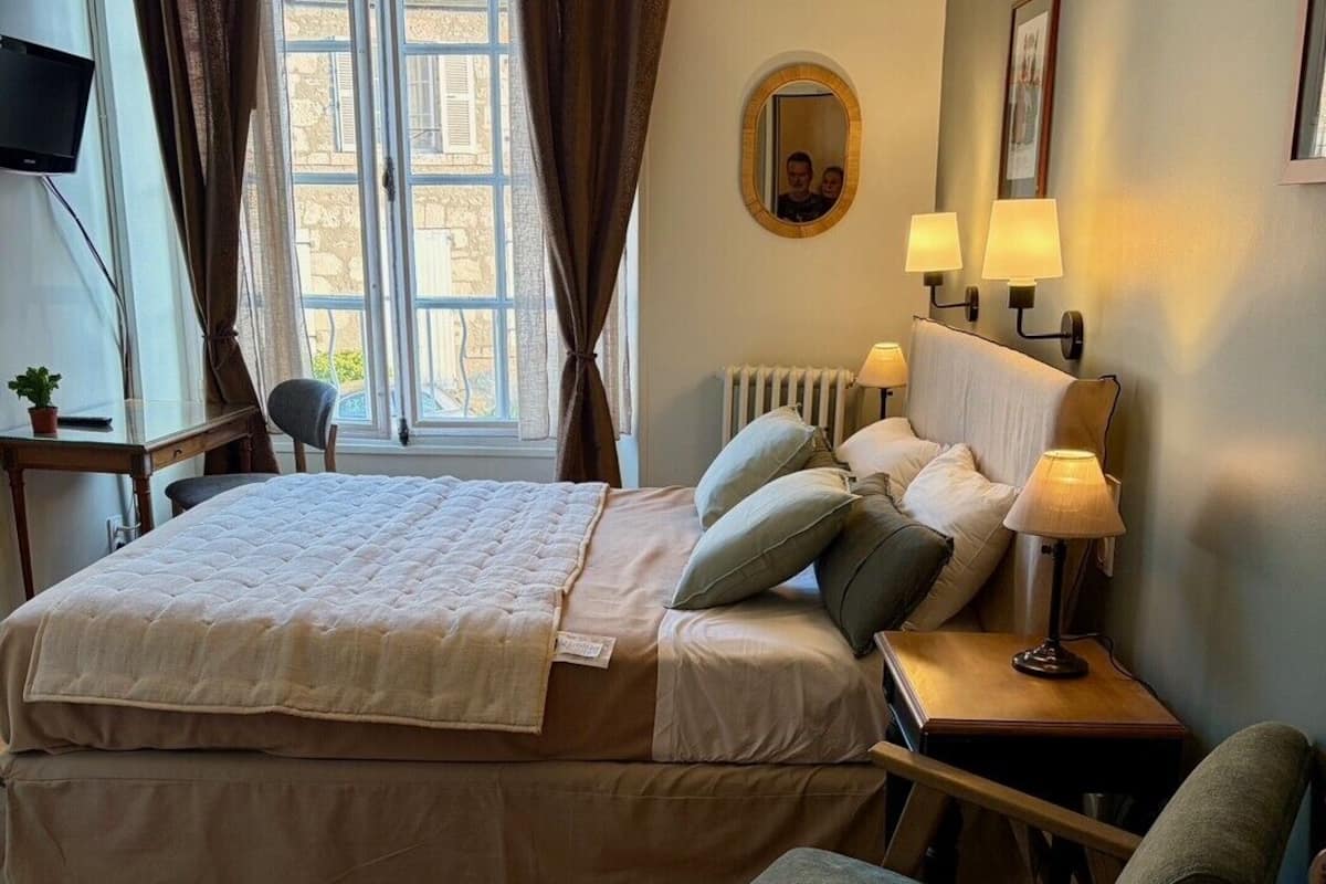 A bedroom with a bed, bedside lamps, a chair, a window with curtains, a radiator, and a wall-mounted mirror.
