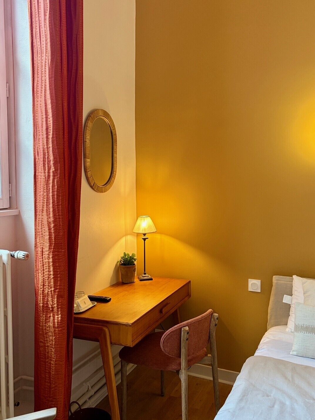 Una habitación de hotel con un escritorio de madera, una silla, una mesita de noche, una lámpara, un espejo y una cama.