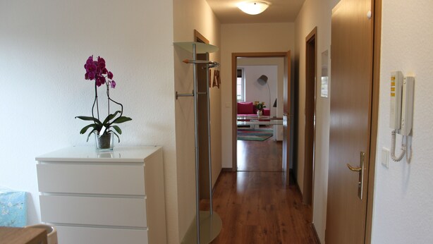 A narrow hallway with wooden flooring, a white dresser with a purple orchid, a glass shelf, and a door with a telephone. - Ferienwohnung Lilie (Lübbenau/Spreewald)