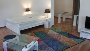 A modern bedroom with a bed, bedside lamps, a glass table, a TV, and a colorful rug. - Ferienwohnung Lilie (Lübbenau/Spreewald)