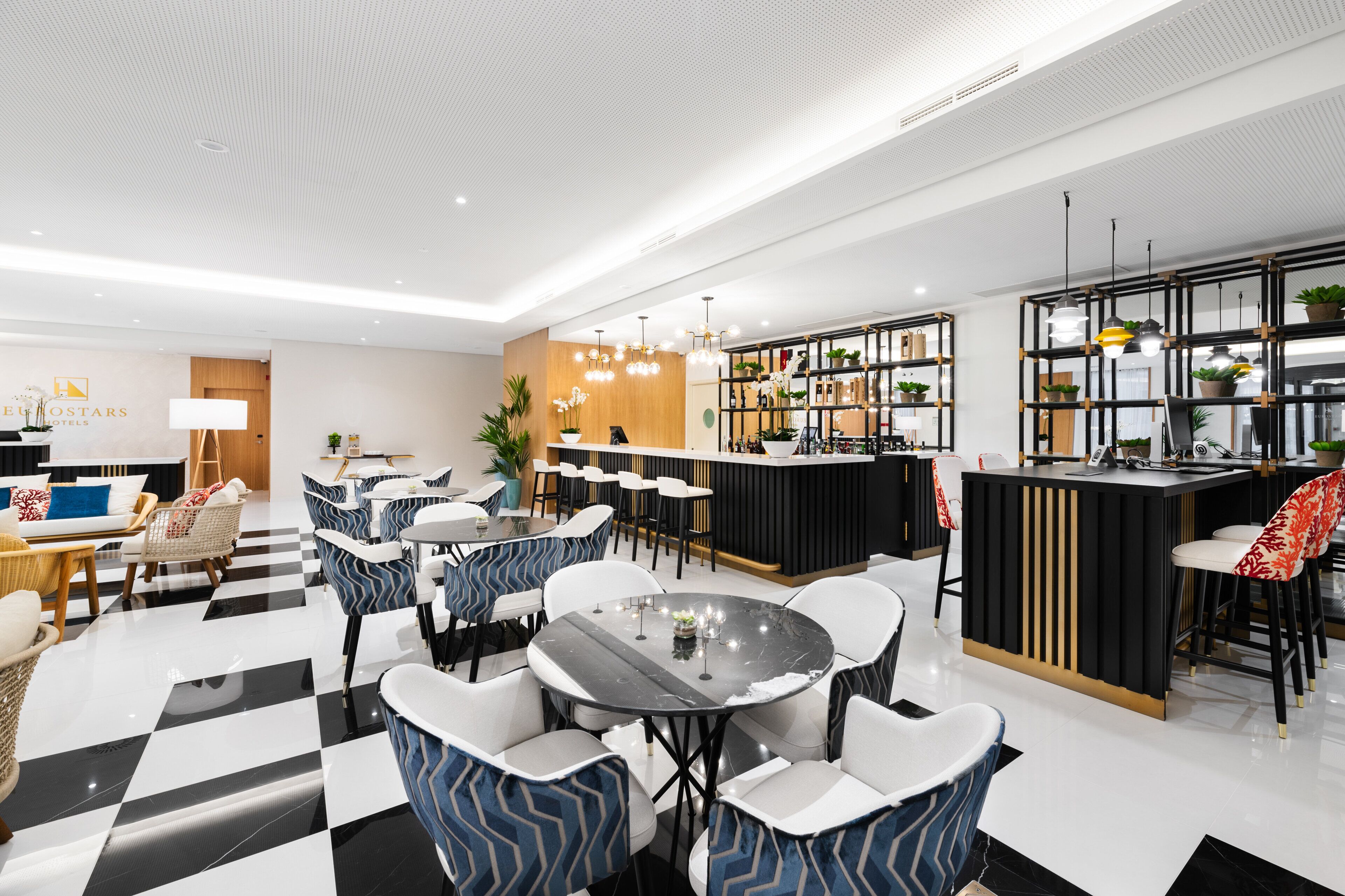 A modern hotel lobby with a checkered floor, seating areas, a reception desk, and a bar area with plants.