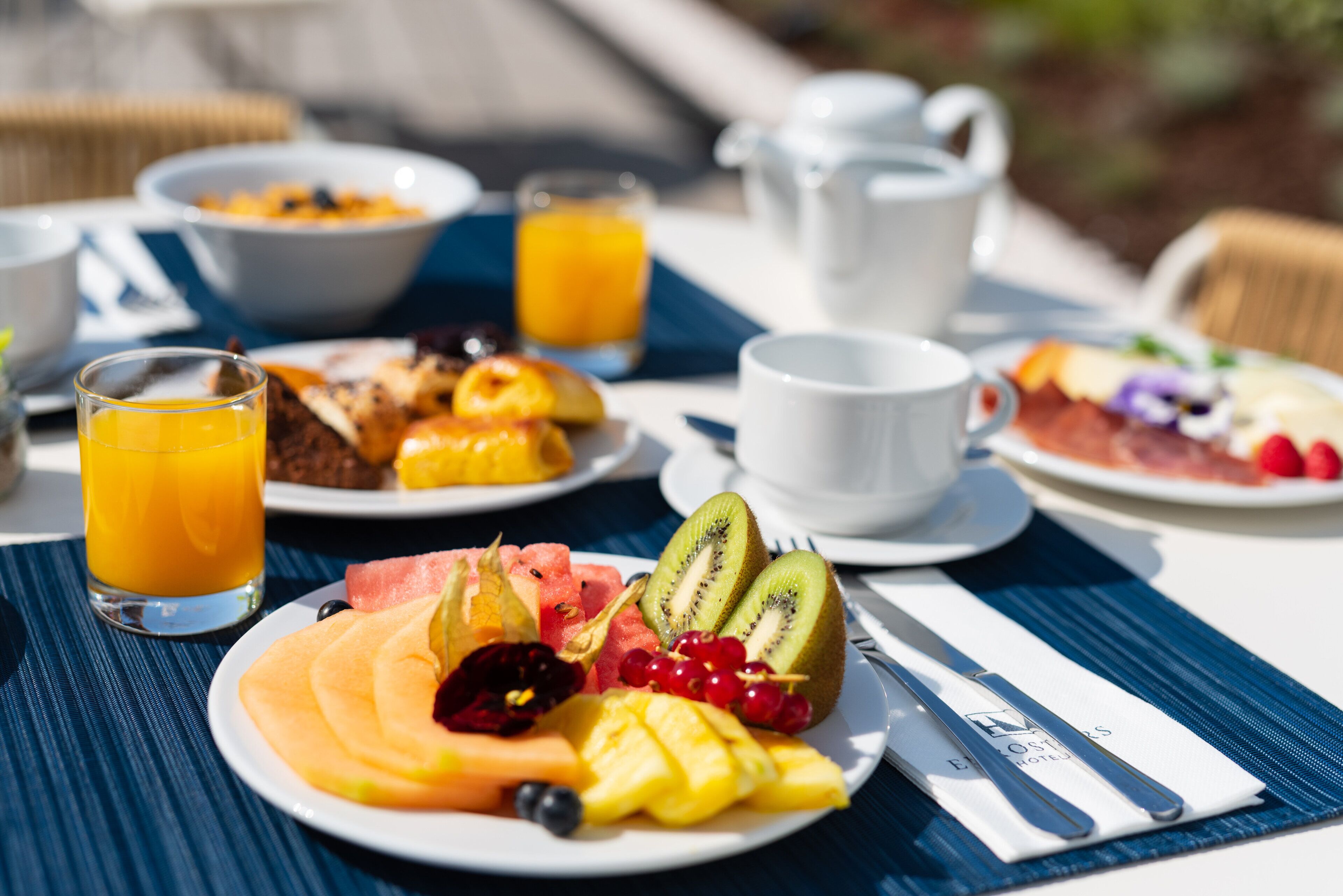 A breakfast setting with plates of fruit, a glass of orange juice, and a white coffee cup on a table.