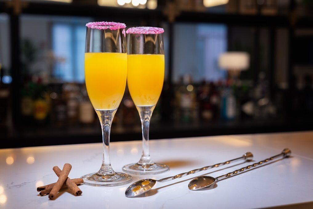 Two glasses of orange juice with pink sugar rims on a bar counter, accompanied by cinnamon sticks and silver spoons.