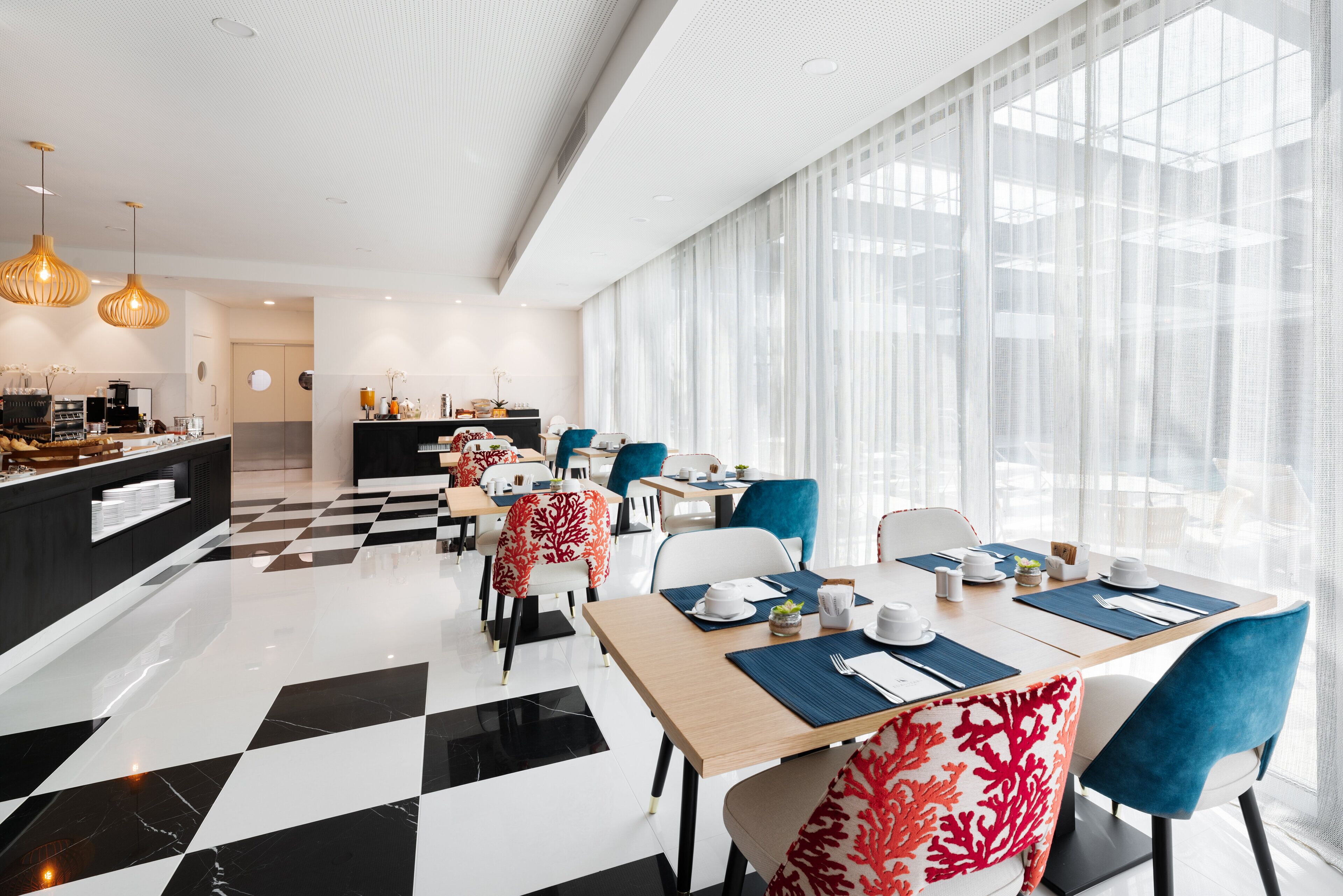 Un comedor de hotel moderno con piso a cuadros, mesas ordenadas con individuales azules y amplias ventanas con cortinas translúcidas.