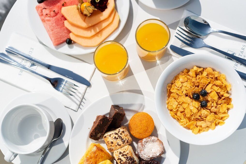 Una mesa de desayuno con platos de comida, vasos de zumo de naranja y productos típicos como cereales y bollería.