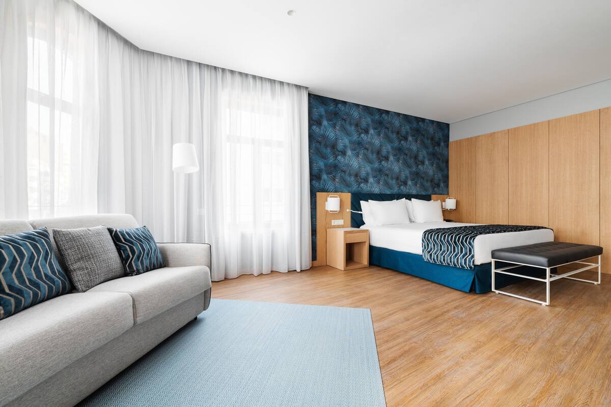 Een moderne hotelkamer met een bank, een bed met een blauw hoofdeinde, een houten vloer en een groot raam met dunne gordijnen.