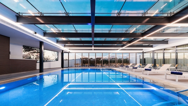 Una piscina interior con techos de vidrio transparente, sillas de descanso y vistas a la ciudad.