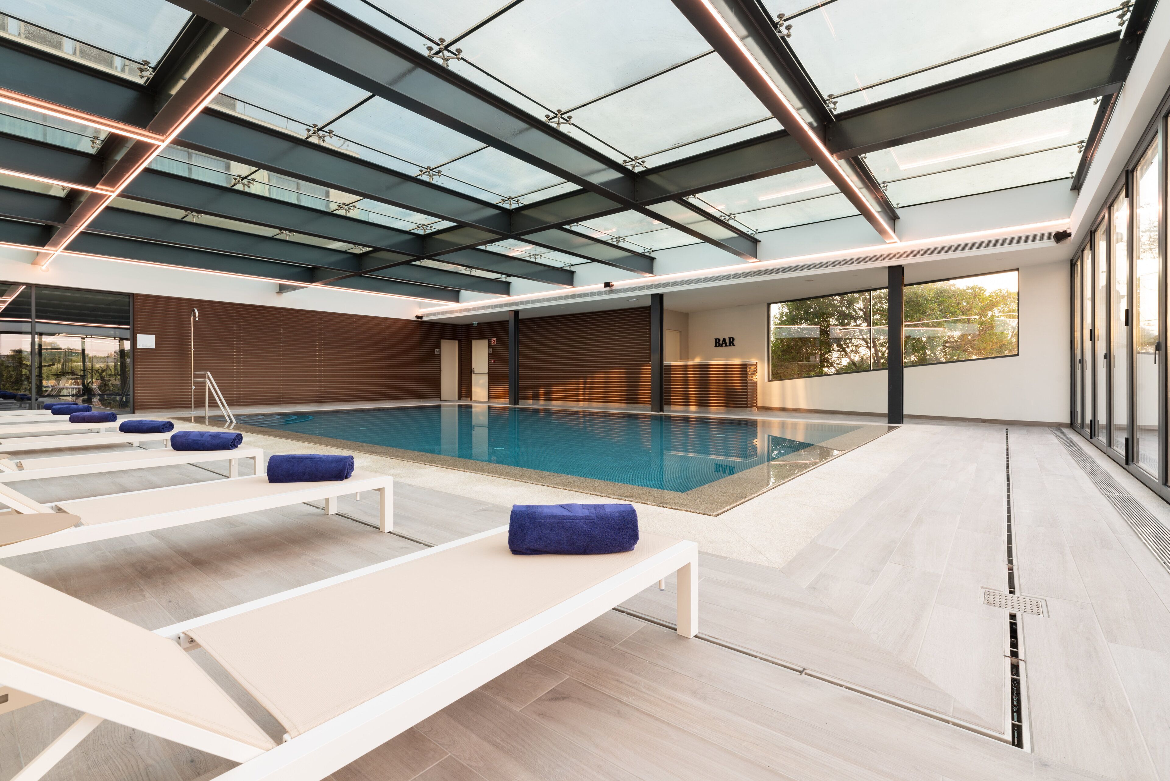 Piscina interior moderna com tetos de vidro, espreguiçadeiras e área de bar.