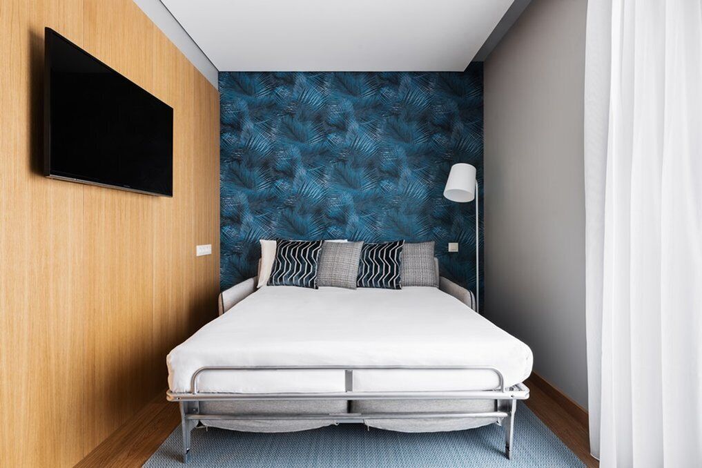 A modern hotel room with a bed, a flat-screen TV mounted on the wall, and a patterned wallpaper.